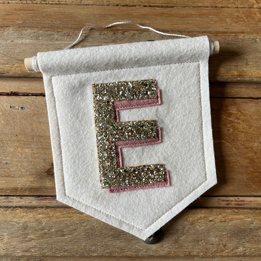 Cream felt initial banner with chunky gold glitter and pink drop shadow effect perfect for kids room decor