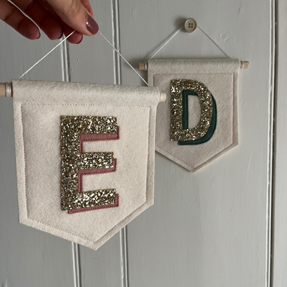 Two personalised initial wall pennants displayed, one hanging on door and the other being held to show perfect for kids room decor.
